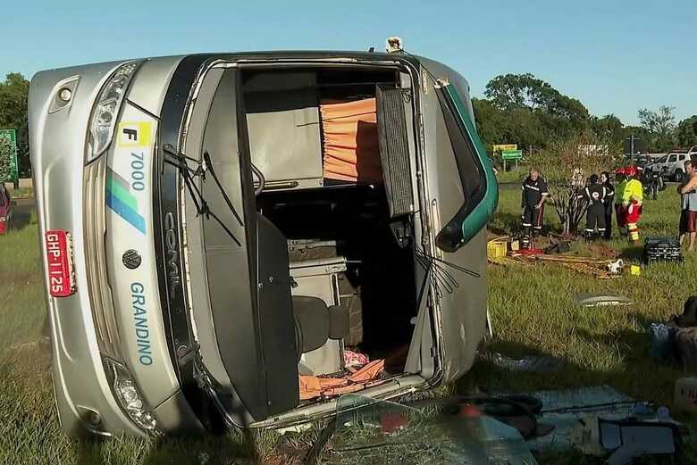 &Ocirc;nibus prata da empresa Grandino tombado de lado em &aacute;rea gramada. Pessoas, incluindo equipe de resgate, est&atilde;o pr&oacute;ximas ao ve&iacute;culo sob c&eacute;u claro. &Ocirc;nibus tombou na pista causando a morte de duas pessoas